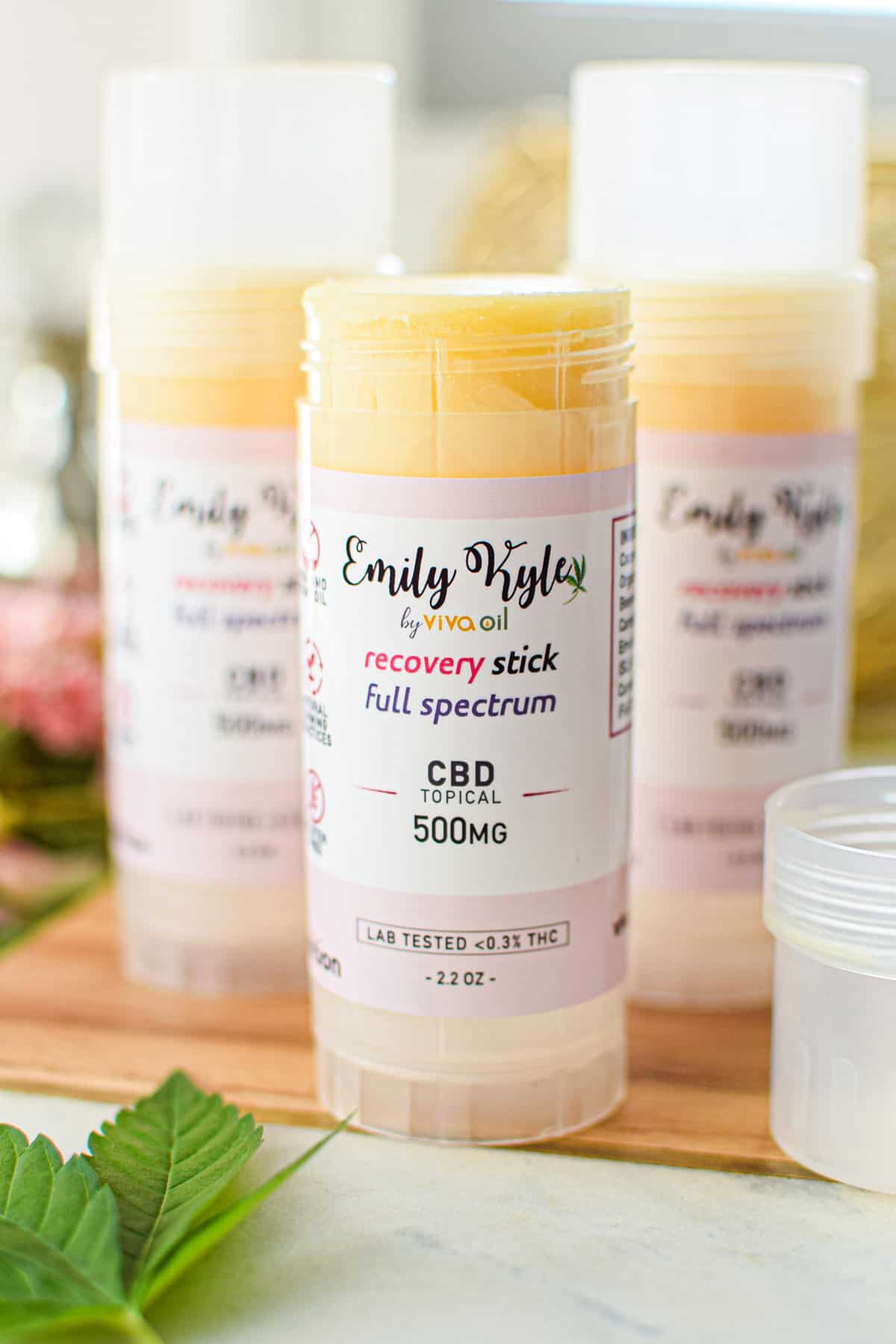 A tube of Emily Kyle's Full-Spectrum CBD Recovery Stick on a white countertop on a cutting board with a cannabis leaf in the back